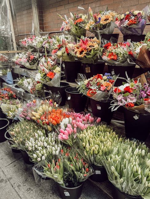 An outdoor display of various fresh floral bouquets and individual flower bunches arranged in black buckets and wrapped in dark and clear plastic, positioned on a paved surface. The bouquets include a