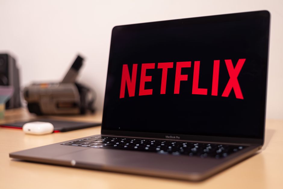 A sleek, open MacBook Pro laptop displaying the Netflix logo on its screen, situated on a wooden desk. The scene includes a white wireless mouse and a black mouse pad in the foreground, with blurred o