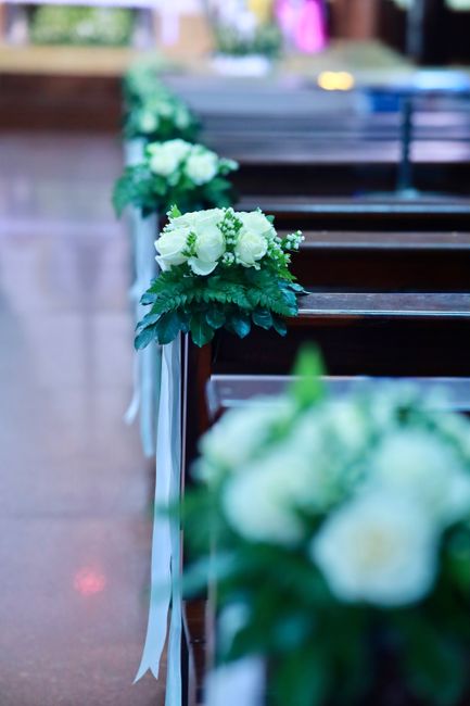 A floral arrangement featuring white roses and hydrangeas, accented with green foliage, is attached to the edge of a polished wooden pew with white ribbon. The bouquet appears freshly crafted, with th